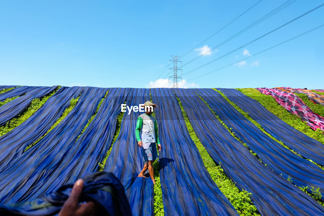 Man drying blue fabrics on grassy field against blue sky during sunny day