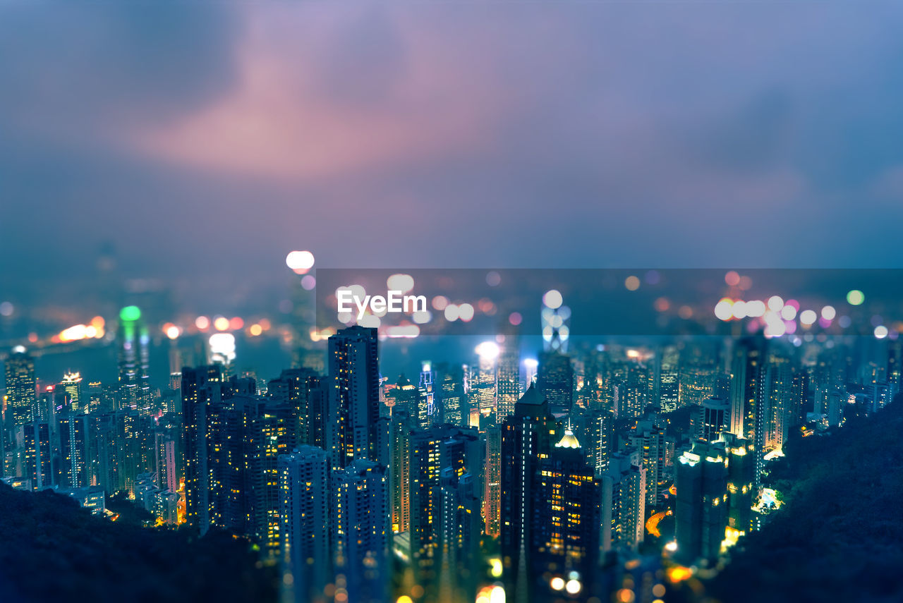 Tilt shift image of illuminated cityscape against cloudy sky at night