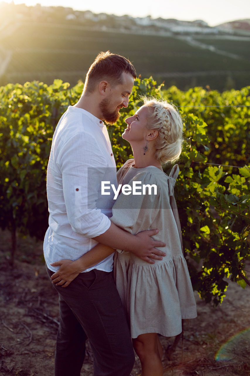 Man and a woman in white clothes stand in a grape field at sunset