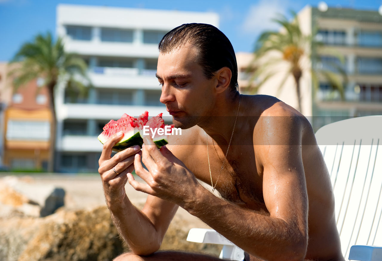 Shirtless man having watermelon against buildings