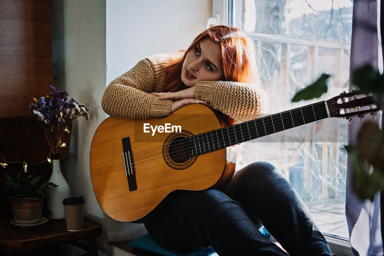 Unaltered candid portrait of young red haired woman in sweater playing acoustic guitar sitting by