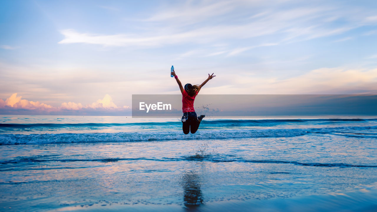 shirtless man with arms outstretched standing at beach against sky during sunset