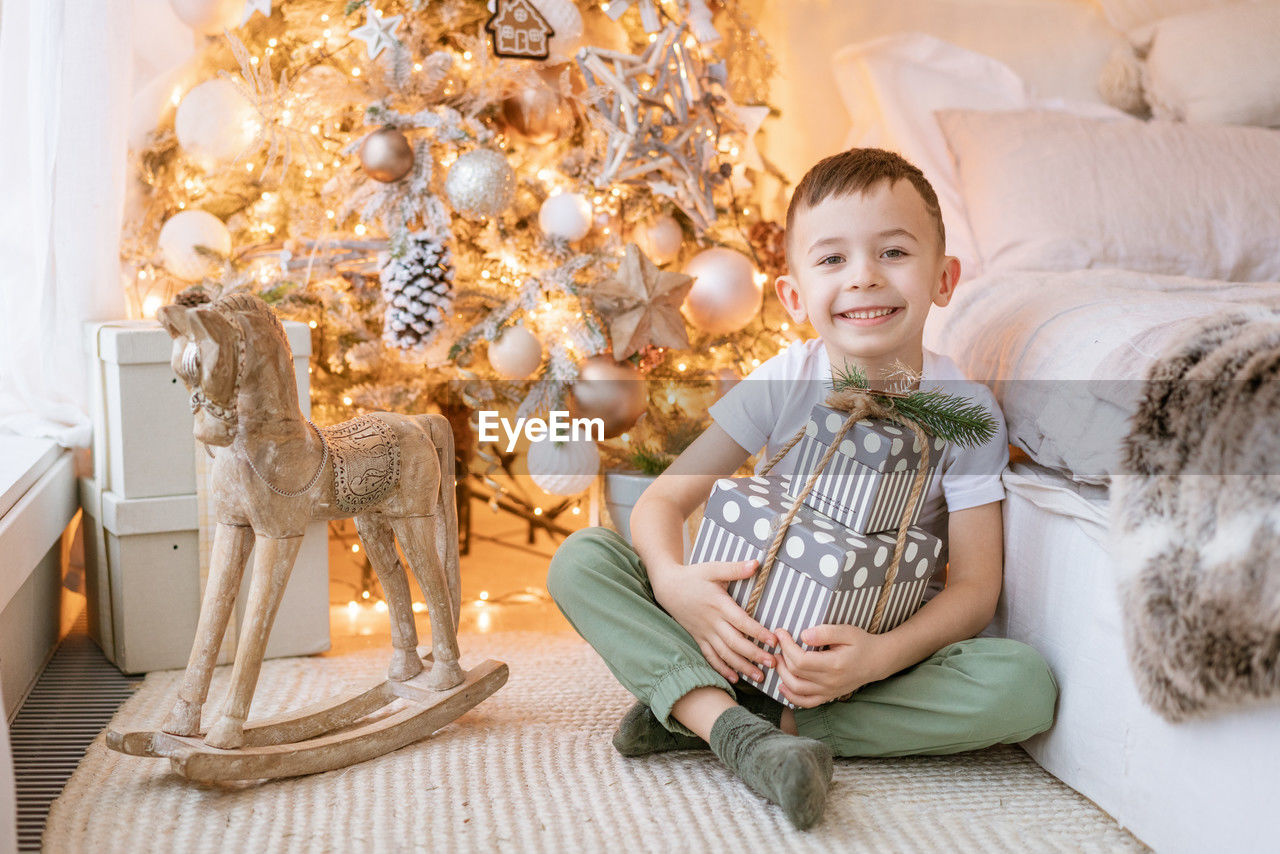 Happy boy sits on floor with gift in his hand by window in front christmas tree