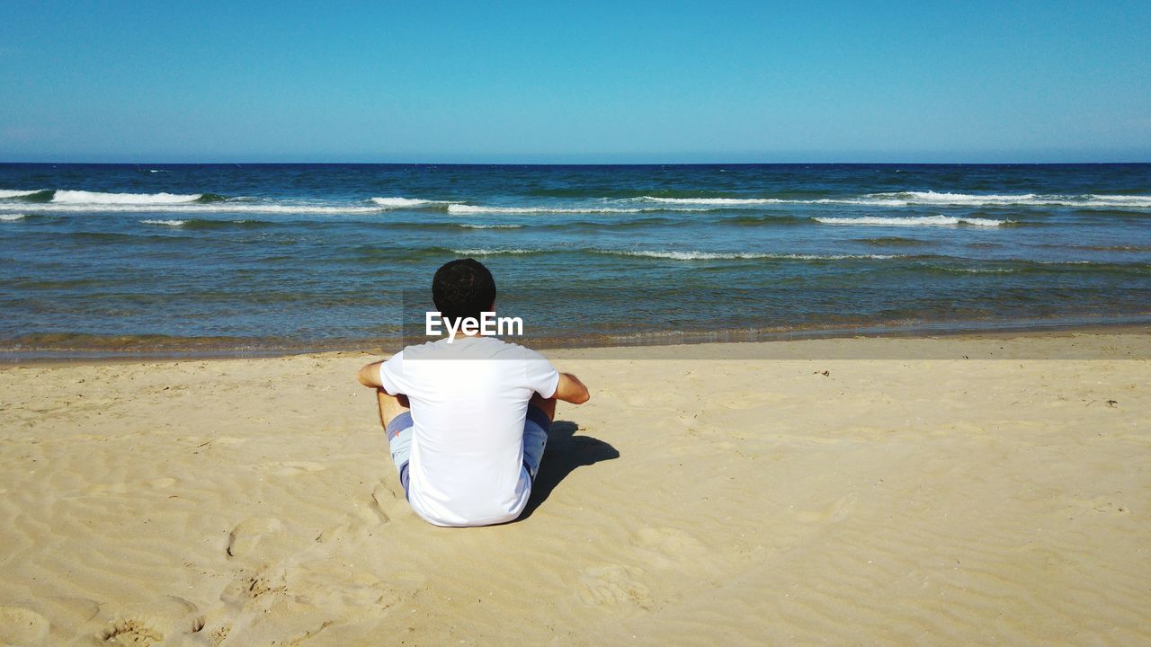 Rear view of man sitting on sand at beach against clear blue sky