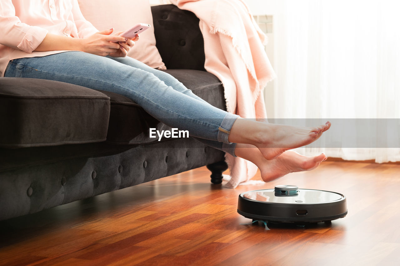 Low section of woman sitting on sofa with robotic vacuum cleaner at home