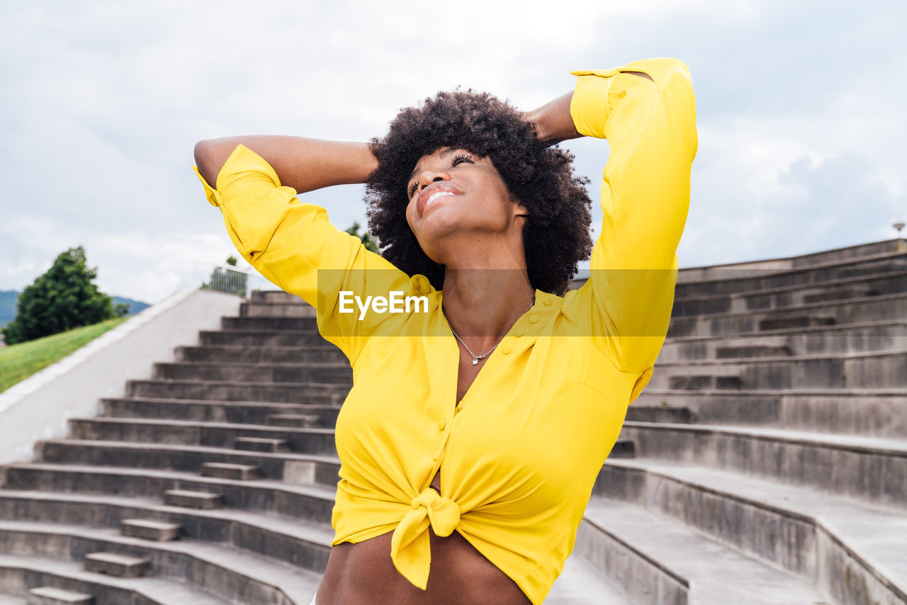 From below optimistic black woman in trendy clothes smiling and dancing with raised arms on sunny day in park