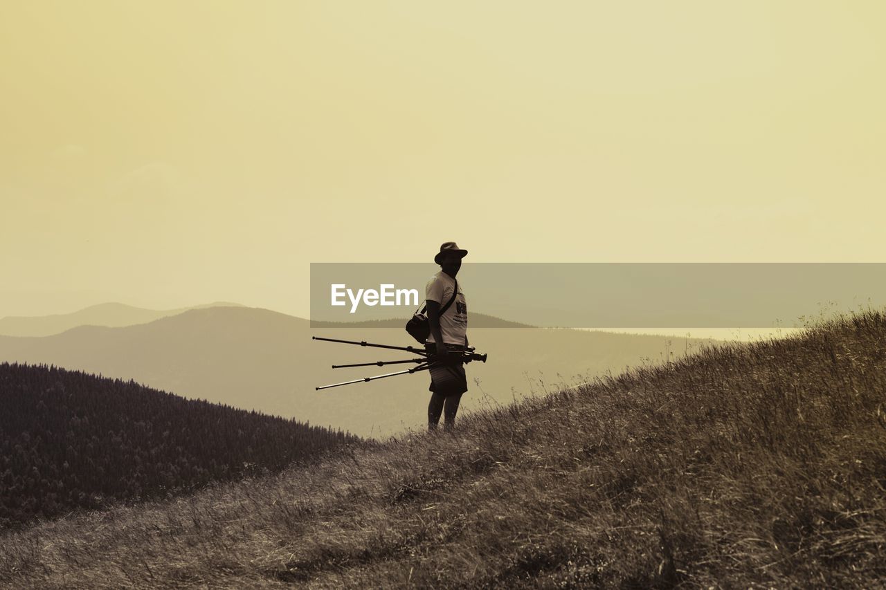 Man holding tripod and camera while standing on field at mountain during sunset