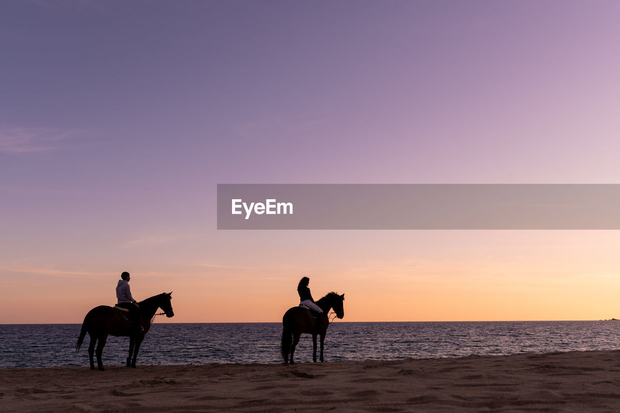 Side view of anonymous couple silhouettes on mares contemplating endless ocean from sandy shore at sundown