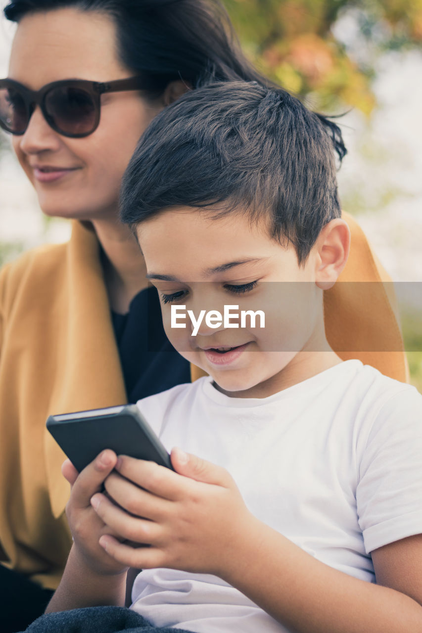 Smiling little boy playing games on smart phone while being in the park with his mother.