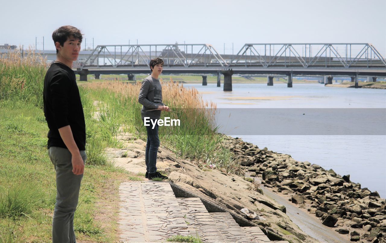 People standing at riverside by bridge against sky