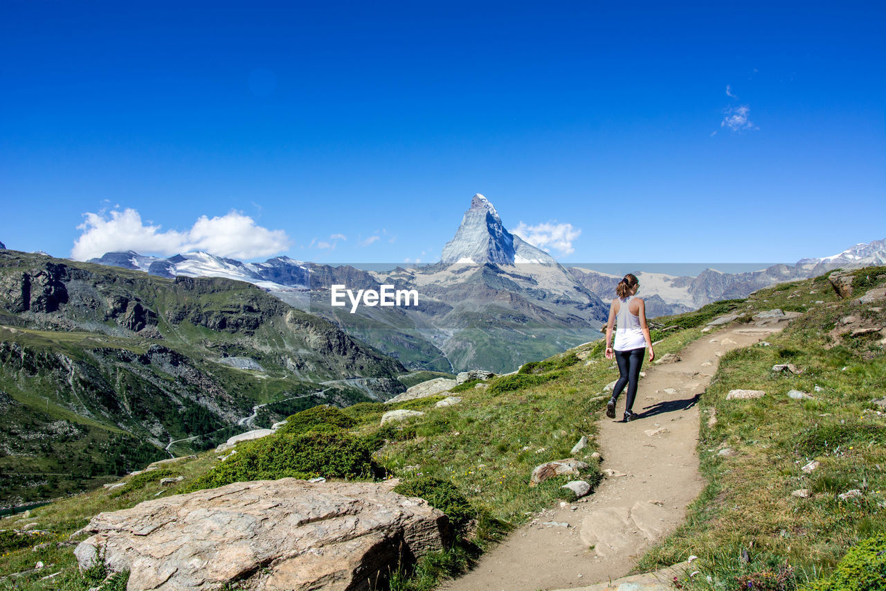 Rear view of woman walking on mountain road against blue sky