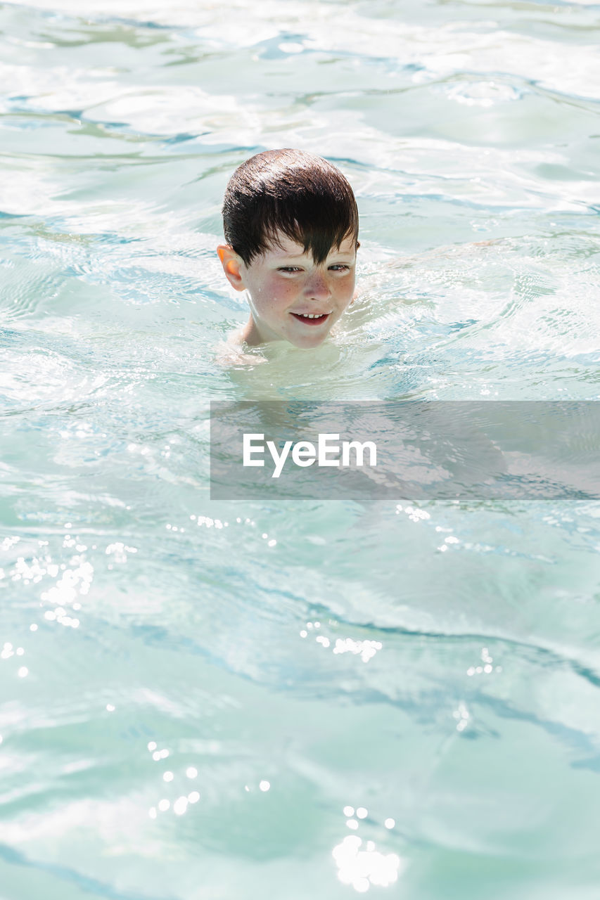 High angle of cute little boy with wet dark hair swimming in outdoor swimming pool during summer holidays