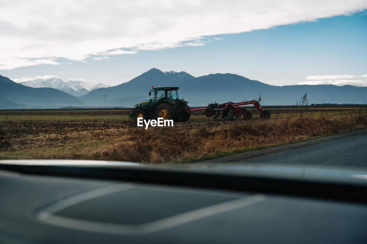 Vehicle on road by agricultural tractor on field against mountain lansdcape and sky during winter