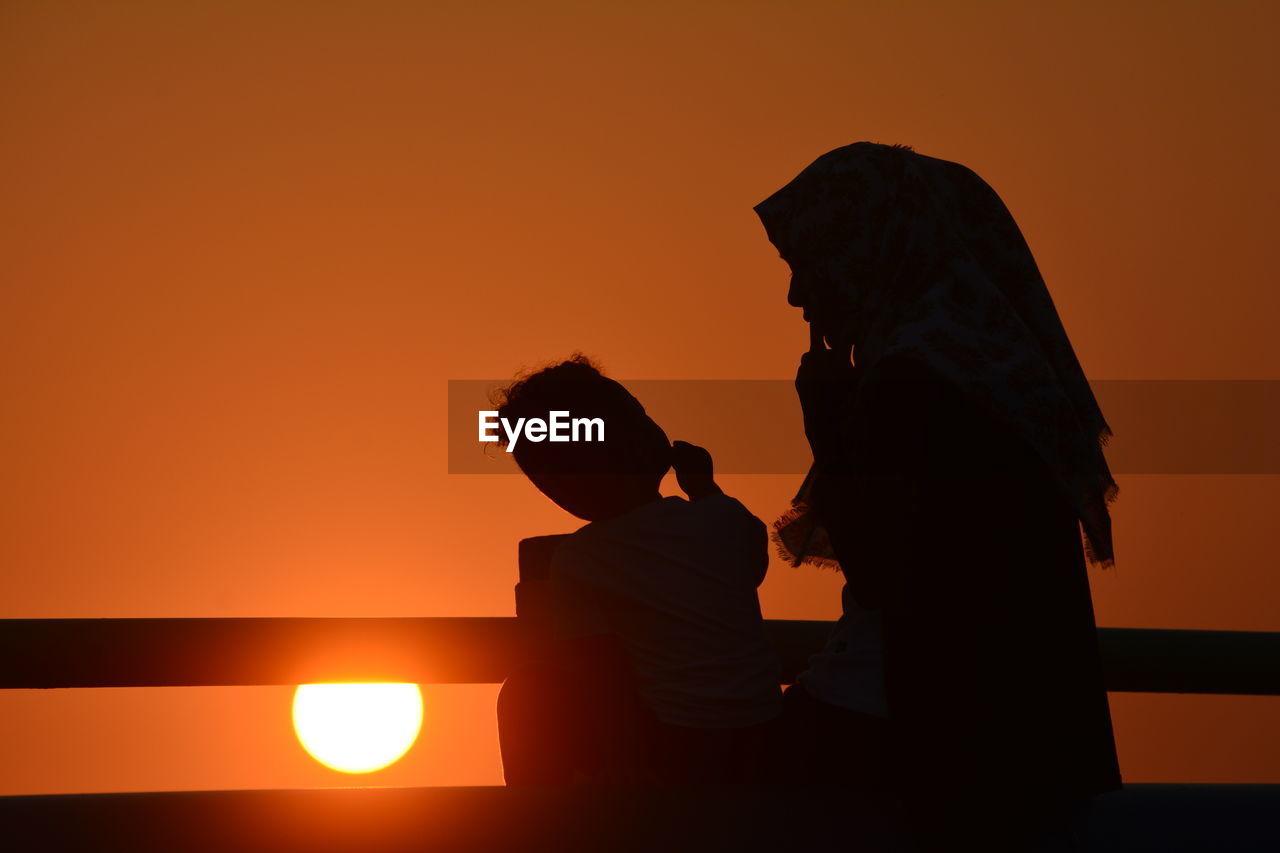 Silhouette mother with son standing by railing against sky during sunset