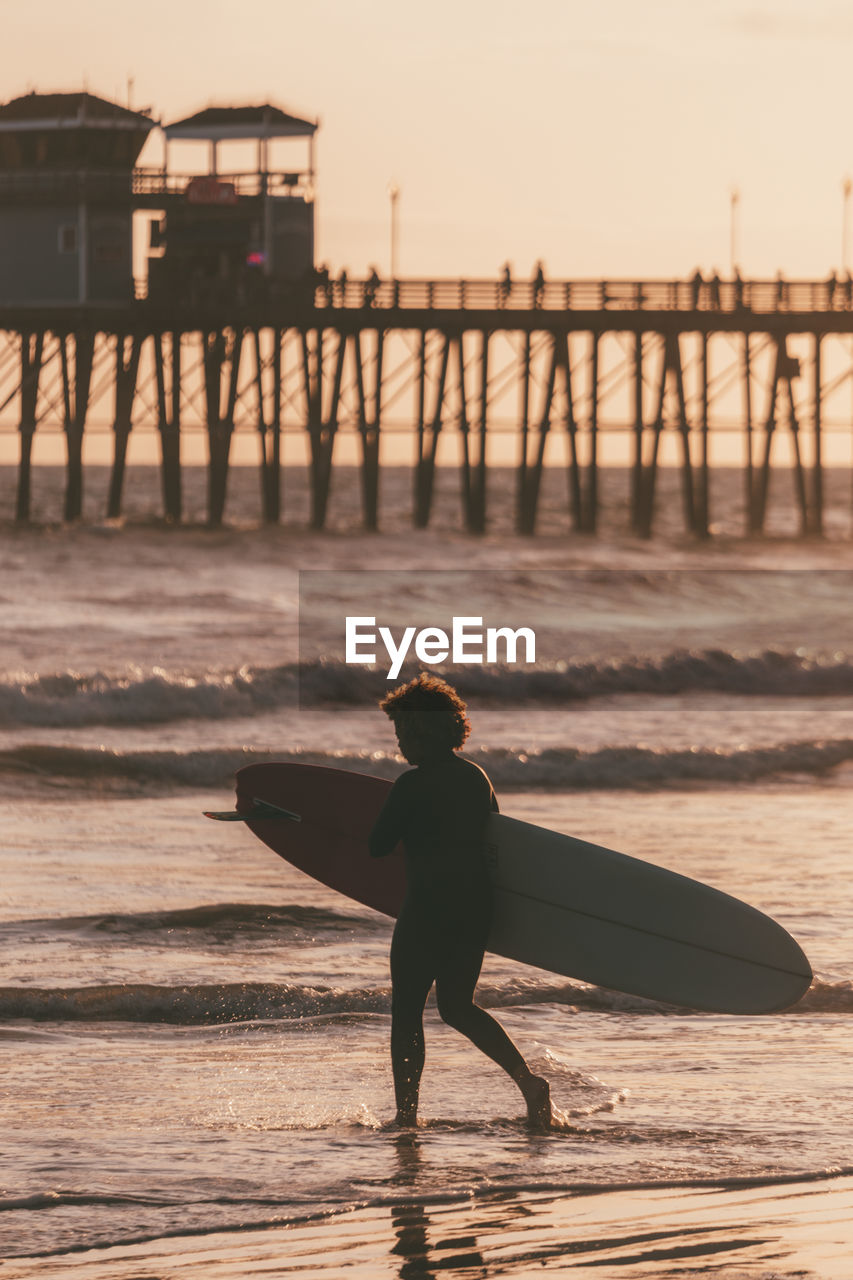 Woman carrying surfboard at sunset by pier