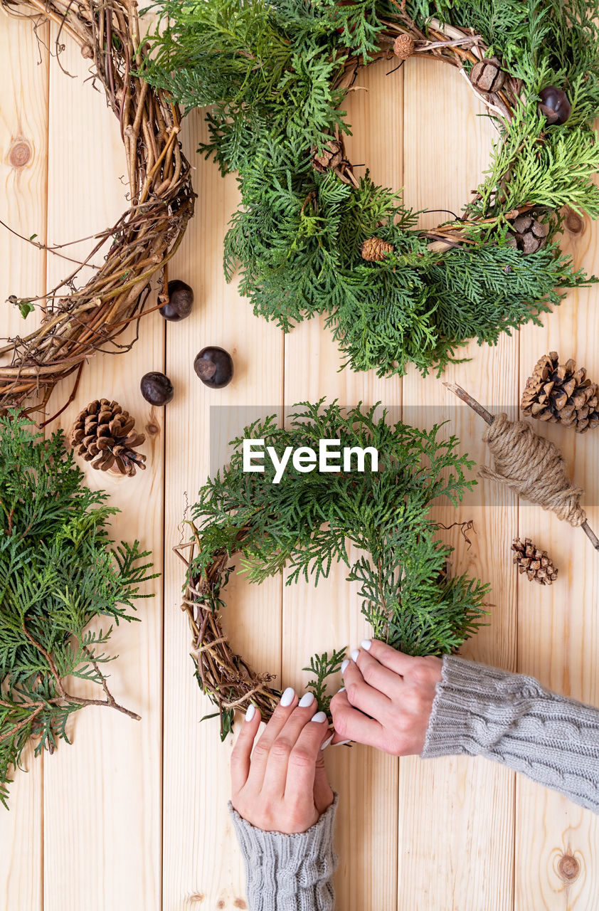  hands making christmas or new year diy wreath with fir tree branches, vines, pine cones top view