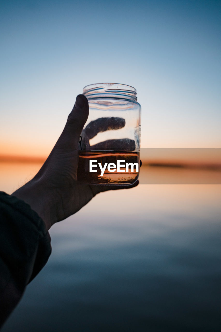 CLOSE-UP OF HAND HOLDING GLASS OF WATER AGAINST SUNSET