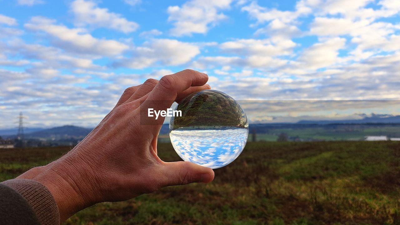 hand, sky, cloud, holding, nature, one person, environment, landscape, water, grass, blue, land, plant, focus on foreground, sphere, outdoors, discovery, personal perspective, day, exploration, travel, adult, close-up, beauty in nature, field, technology