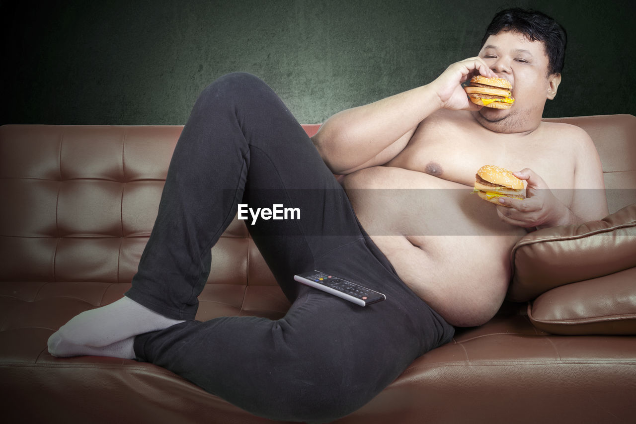 Shirtless man eating burger while leaning on sofa at home