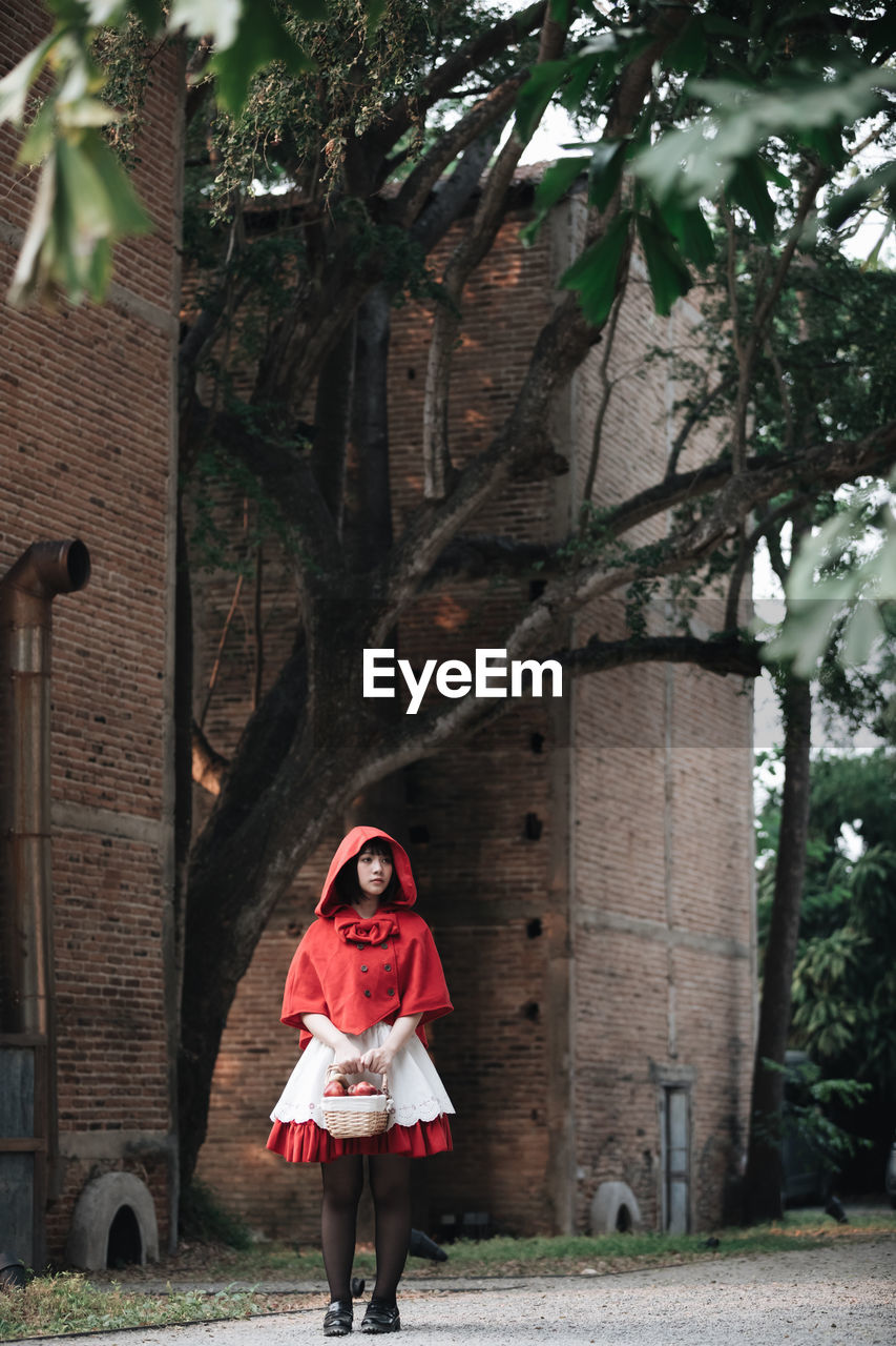 Full length of woman in little red riding hood costume holding basket while standing against building