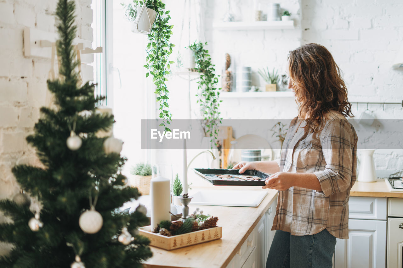 Attractive smiling woman with curly hair in plaid shirt bakes cookies at bright kitchen at home