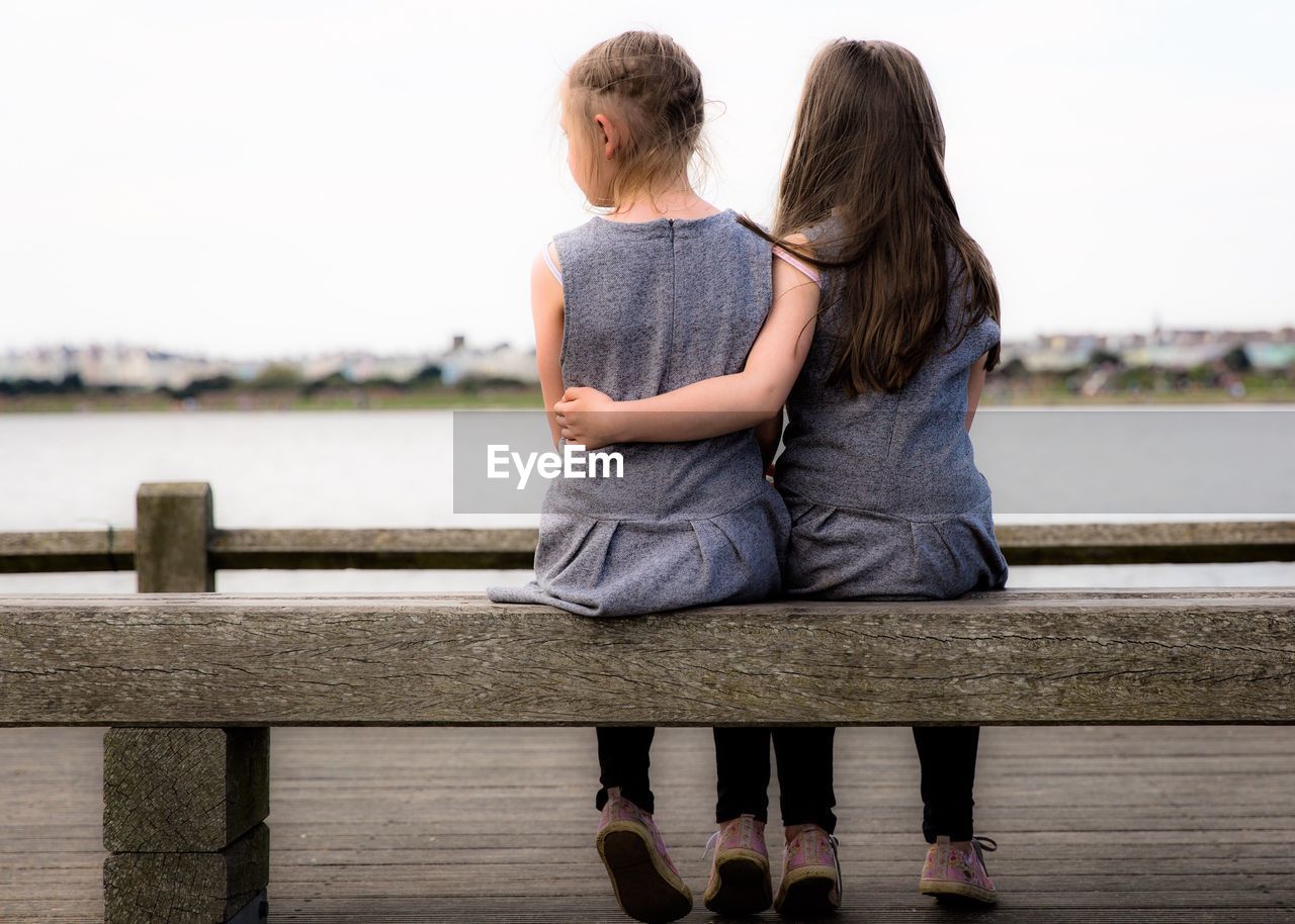 Full length rear view of girls sitting on pier against clear sky