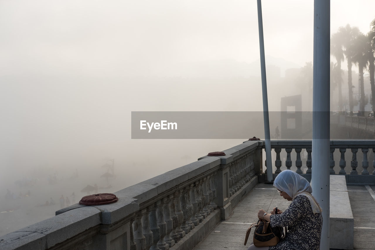 WOMAN SITTING ON FOOTBRIDGE IN FOGGY WEATHER