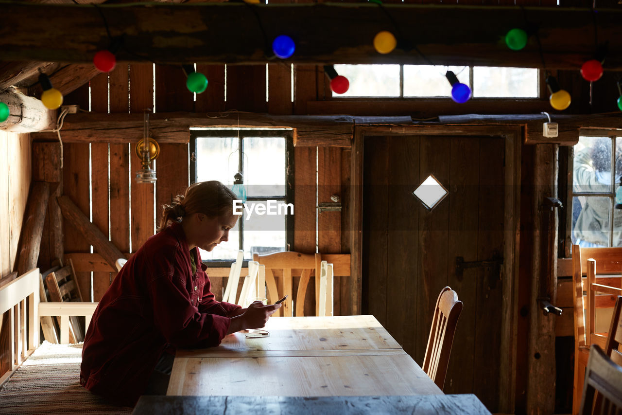 Side view of young woman using smart phone while sitting at table in log cabin