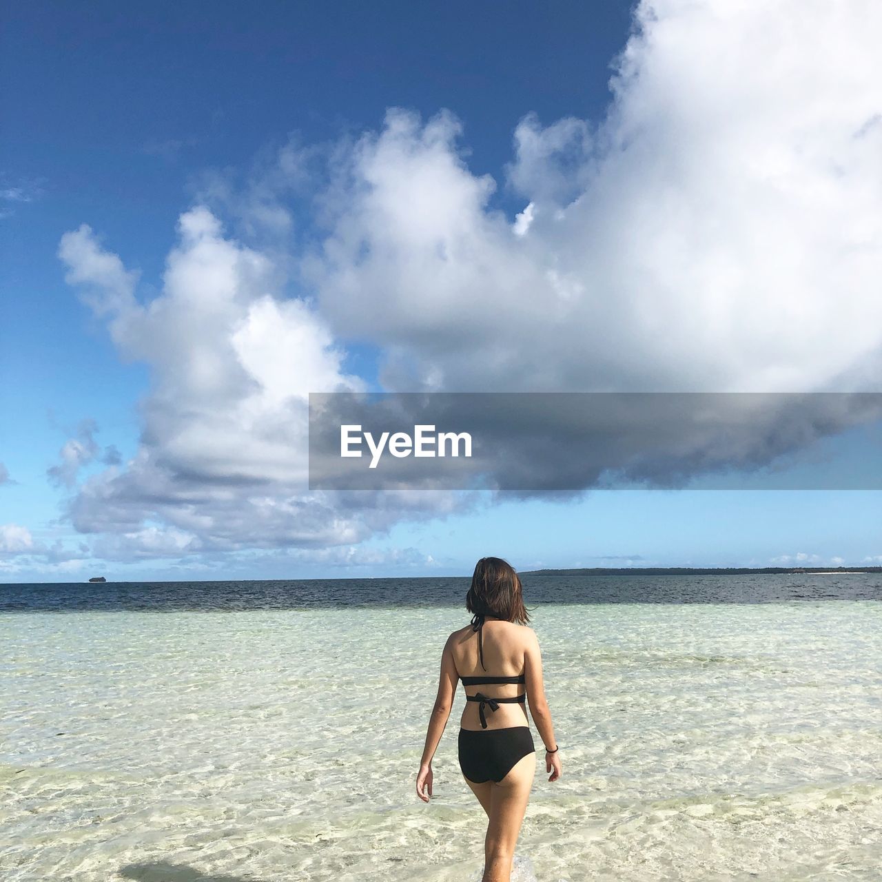 Rear view of woman in bikini walking at beach against cloudy sky