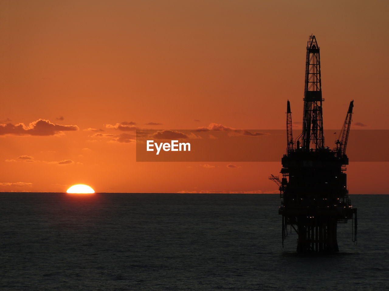 Silhouette oil industry in sea against sky during sunset