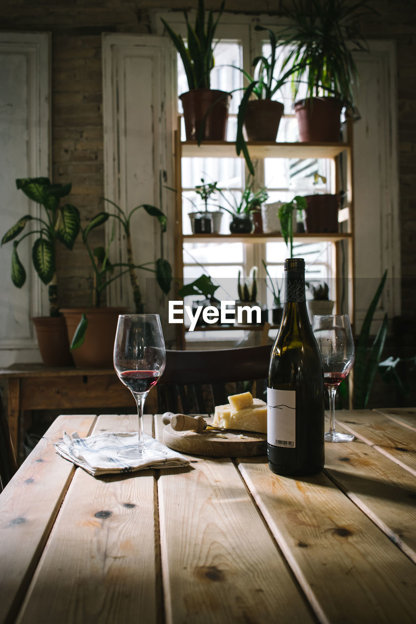 Open bottle and glasses with red wine placed near cheese on wooden table in rustic restaurant with potted green plants on window