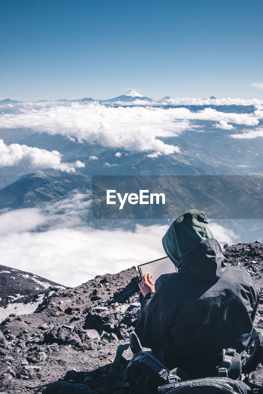 Rear view of man drawing while sitting on mountain against sky