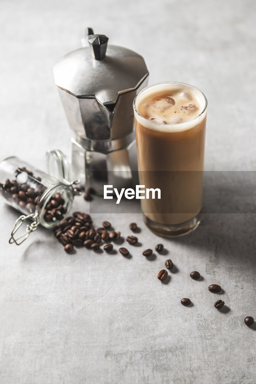 Close-up high angle view of iced coffee cup, moka pot and spilled coffee beans on grey concret table