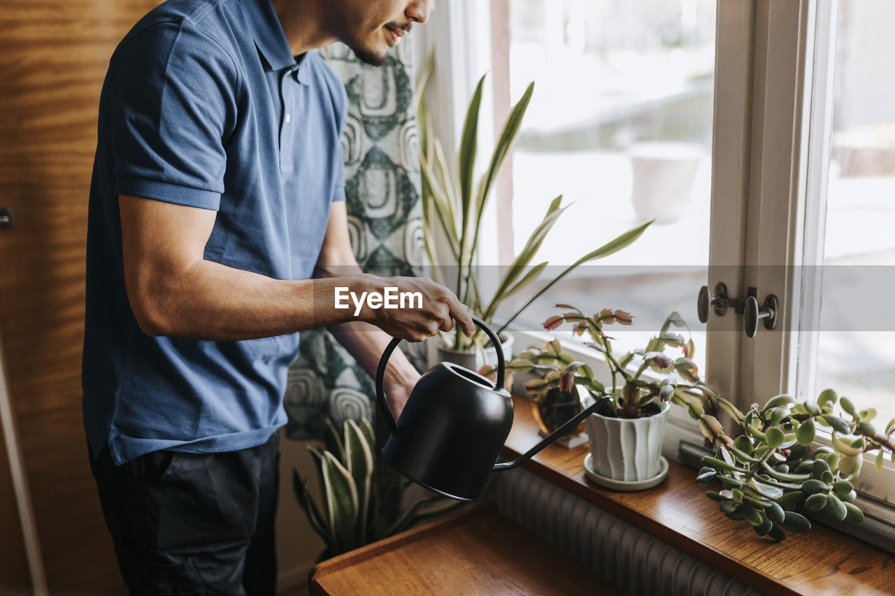 Midsection of male caregiver watering plants on window sill at home