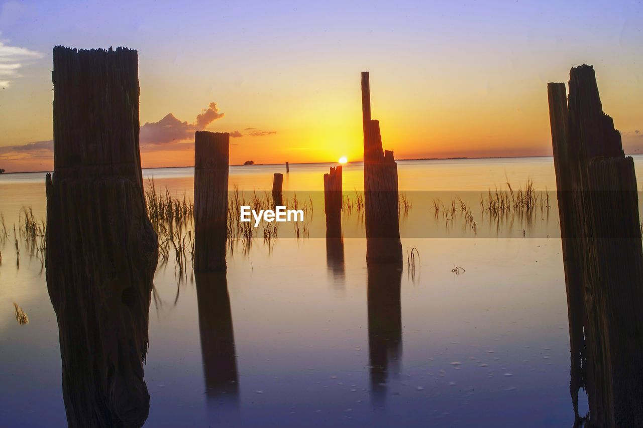 Wooden posts in sea against sky during sunset