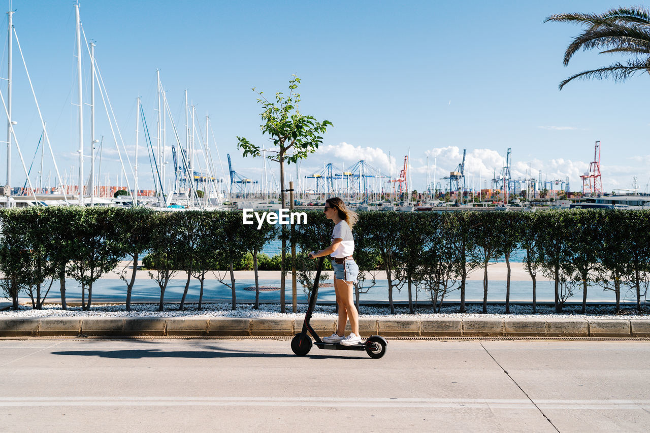 Full body side view of young female in casual outfit riding electric scooter on paved road near urban embankment in sunny summer day