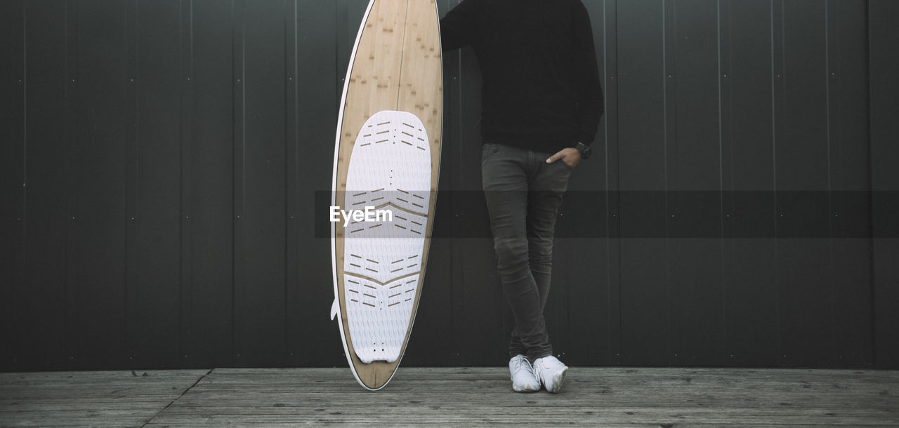 Low section of man with surfboard standing against wall