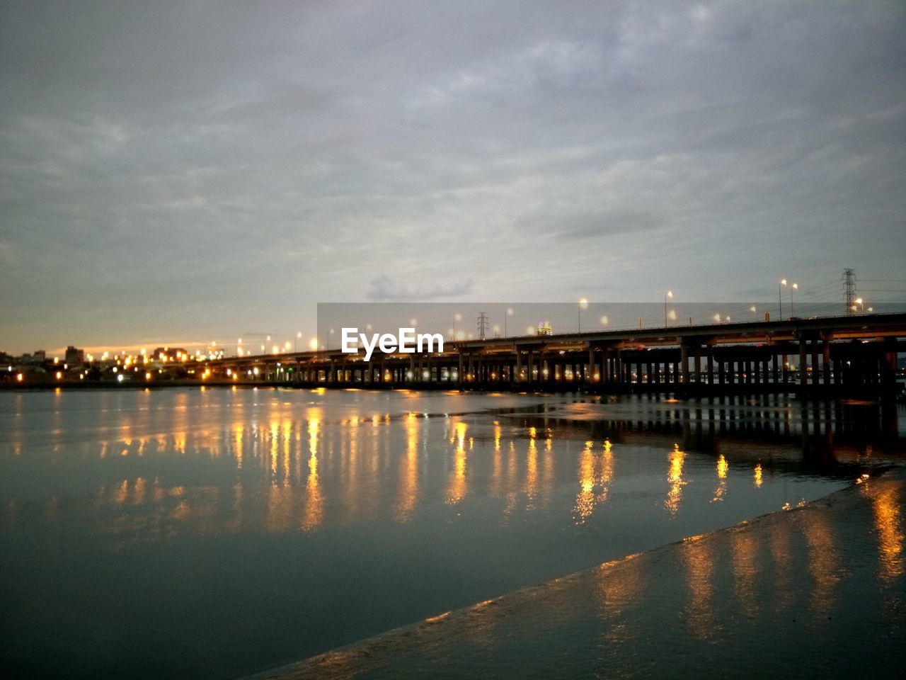 ILLUMINATED BRIDGE OVER RIVER BY CITY AGAINST SKY