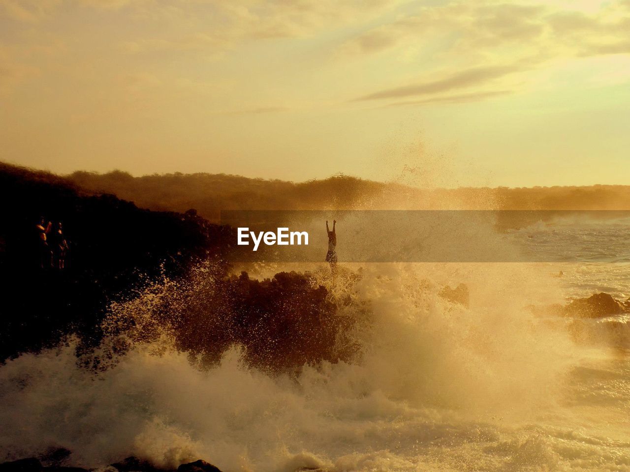 Waves splashing at rocky shore against cloudy sky during sunset