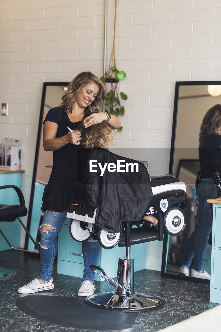 Full length of smiling hairdresser cutting girl's hair in salon