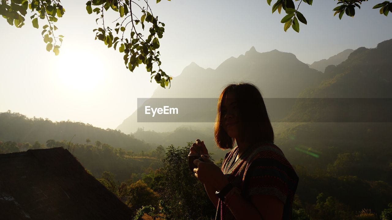 Portrait of woman holding coffee cup by mountains against sky