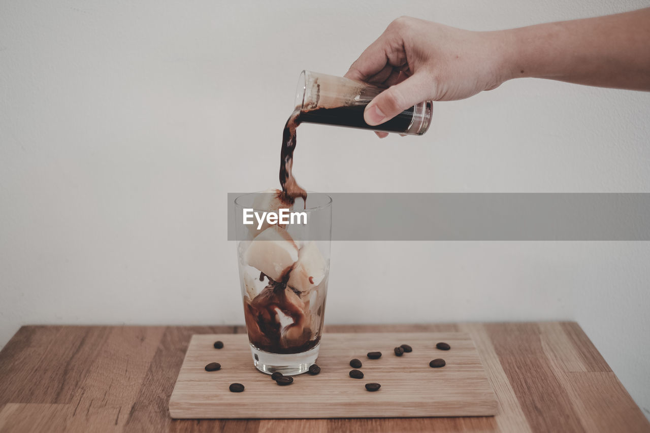 Cropped hand of person pouring drink in drinking glass on table against wall