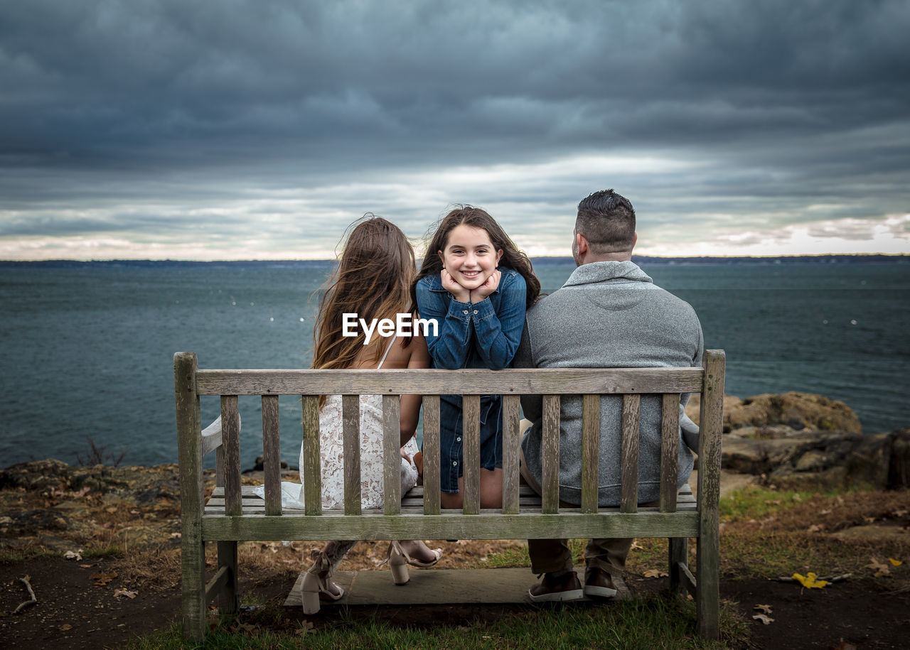 REAR VIEW OF COUPLE SITTING ON SHORE AGAINST SEA