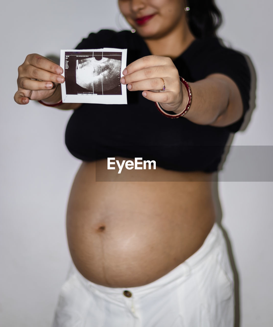 adult, studio shot, trunk, women, one person, holding, indoors, portrait, pregnant, clothing, female, standing, person, hand, smiling, anticipation, happiness, young adult, arm, gray background, parent, gray, white background, beginnings, finger, emotion, technology, looking at camera, front view, life events, waist up, photo shoot, limb, positive emotion, lifestyles, child, white