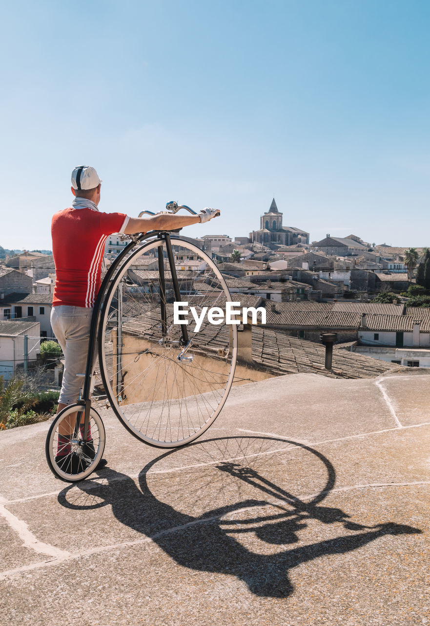 From below back view of unrecognizable man in casual clothes looking away and enjoying view of old city under cloudless blue sky while standing with penny farthing bike on street