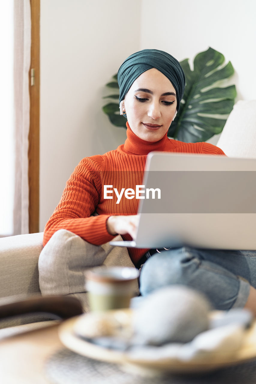 Muslim female in stylish turtleneck and headscarf using laptop and listening to music on sofa on sunny day at home