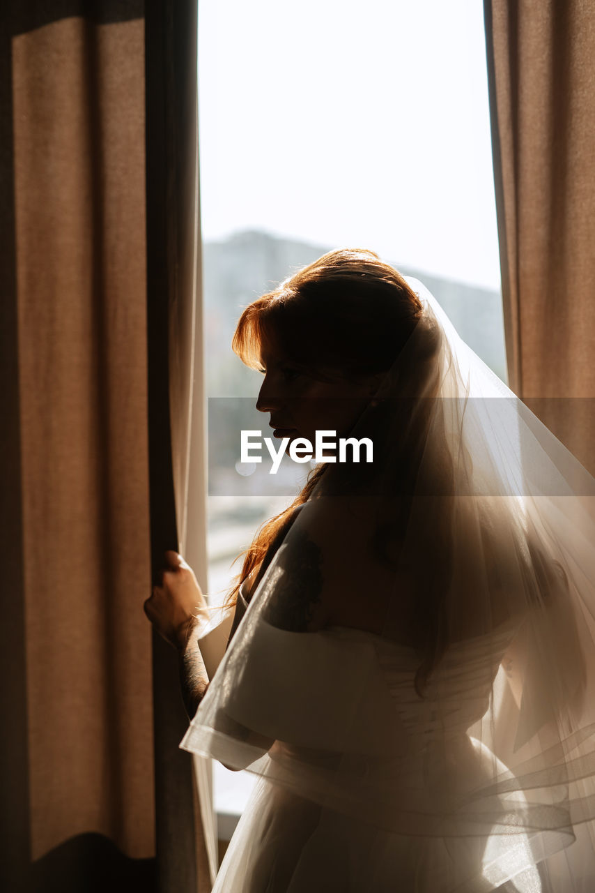 one person, wedding dress, bride, adult, window, indoors, curtain, women, dress, female, side view, standing, looking, young adult, veil, three quarter length, contemplation, sunlight, holding, day, anticipation, lifestyles, waist up, hairstyle, looking through window, person, emotion, clothing, looking down, long hair, brown hair, nature, back lit, gown, photo shoot