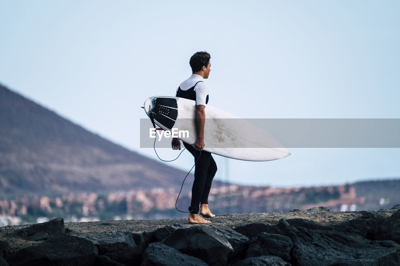 Teenage boy holding surfboard while standing on rock against clear sky