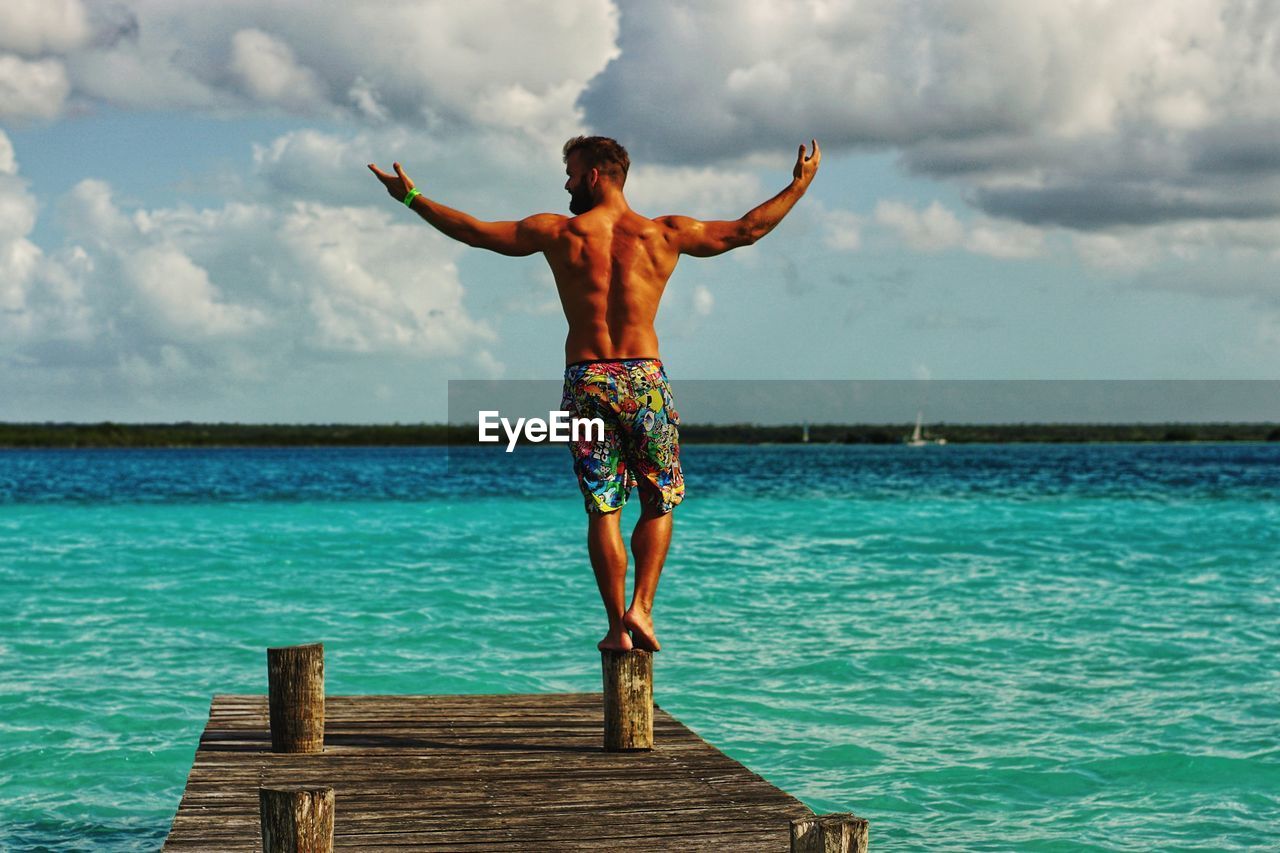 Rear view of shirtless man posing while standing on pier against sky