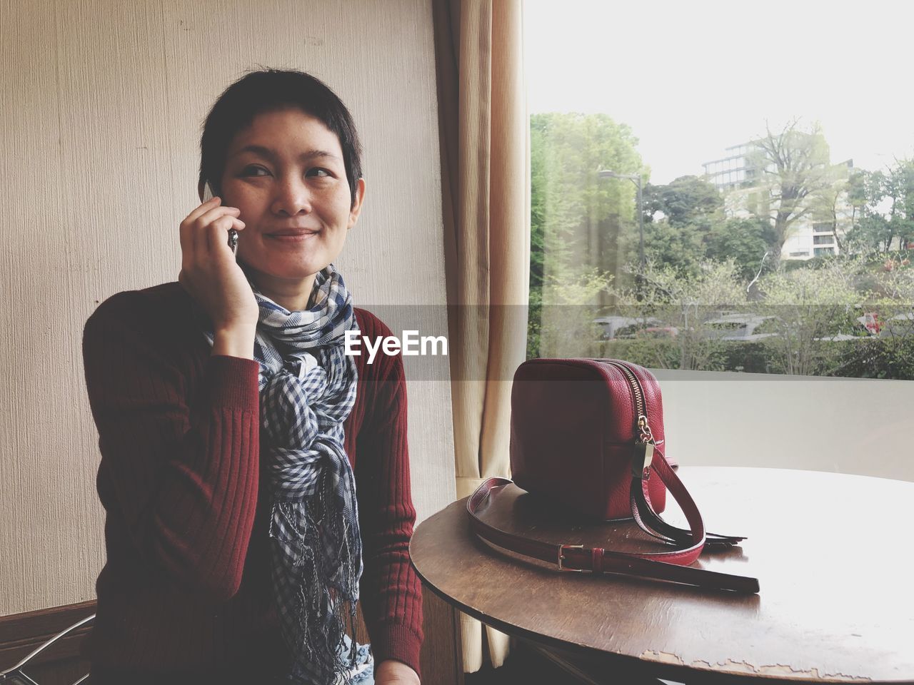 PORTRAIT OF YOUNG WOMAN USING SMART PHONE WHILE STANDING ON WINDOW